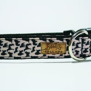 May include: A black and pink dog collar with a silver D-ring and a black buckle. The collar has a repeating pattern of black silhouettes of dogs on a pink background. The collar has a brown leather tag that says "Bowes Collars".