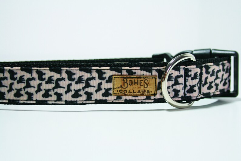 May include: A black and pink dog collar with a silver D-ring and a black buckle. The collar has a repeating pattern of black silhouettes of dogs on a pink background. The collar has a brown leather tag that says "Bowes Collars".