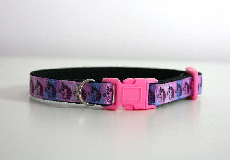 Pu&ograve; includere: Un collare per animali domestici con un motivo ripetuto di teste di unicorno rosa, viola e blu. Il collare ha una base nera, un anello a D in metallo argentato e una fibbia in plastica rosa brillante.