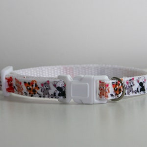 May include: White cat collar with a colorful pattern of cartoon cats. The collar has a white plastic buckle and a silver metal ring.