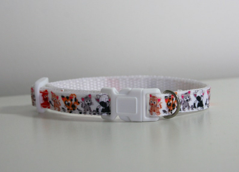 May include: White cat collar with a colorful pattern of cartoon cats. The collar has a white plastic buckle and a silver metal ring.