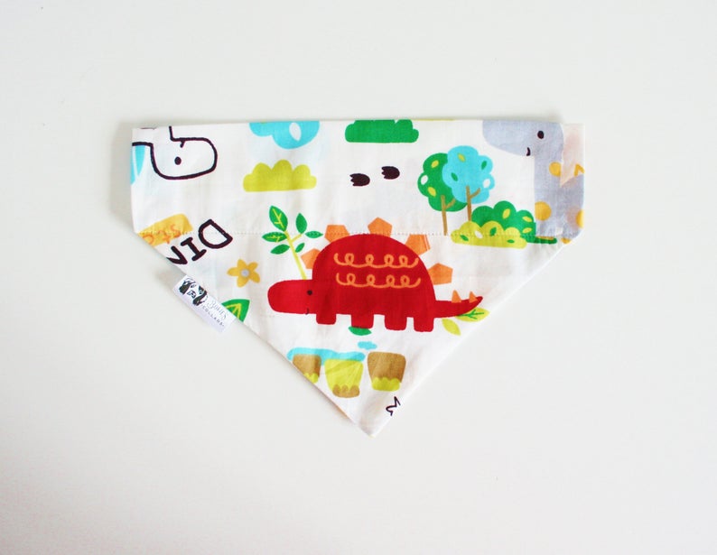 May include: A white triangular bandana with a dinosaur print. The bandana features red, green, yellow, blue, and gray dinosaurs, trees, and clouds. The text "DIN" is visible on the bandana.