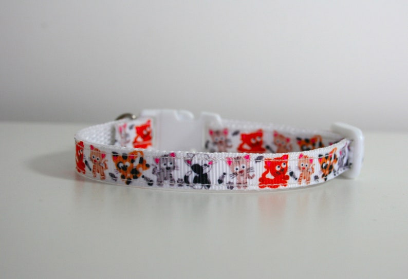 May include: White cat collar with a colorful pattern of cartoon cats. The collar has a plastic buckle.
