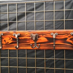 May include: A rectangular wall-mounted coat rack featuring a burnt orange and dark brown wood grain finish. The rack includes four metal hooks with scrollwork tops and one central metal hook shaped like a ram head with curved horns.