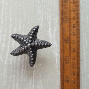 May include: A dark grey, textured starfish-shaped cabinet knob with a bumpy surface. The knob is positioned next to a wooden ruler that shows the object spans approximately 6.5 cm in width and height, providing a clear scale for the item.