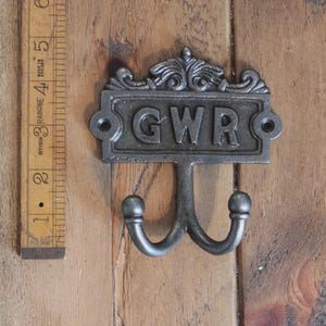 May include: A dark grey metal wall hook featuring the letters GWR embossed on a rectangular plate with decorative scrollwork at the top and two curved hooks at the bottom, positioned next to a wooden ruler measuring approximately 14 cm.