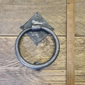 May include: A dark grey metal door knocker, featuring a diamond-shaped backplate and a large, circular handle. The knocker has a distressed finish, giving it an antique appearance. It is mounted on a wooden surface. The knocker is made of metal.