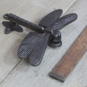 May include: A dark grey cast iron dragonfly door knocker. The dragonfly features detailed wings and body. A wooden ruler is next to the door knocker. The ruler is approximately 15 cm long.