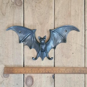 May include: A dark grey metal bat wall decoration with outstretched wings. The bat has detailed wing and body features. A wooden ruler is below the bat, and the background is wooden planks.