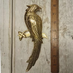 May include: A brass bird sculpture perched on a branch, with detailed feather and wing designs. The bird is approximately 20 cm long, with a ruler alongside for scale. The sculpture has a vintage aesthetic.