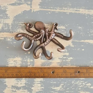 May include: Bronze octopus wall hook with eight curved tentacles. The octopus has a textured surface and is mounted on a distressed, light blue and grey wooden background. A wooden ruler is below the octopus.