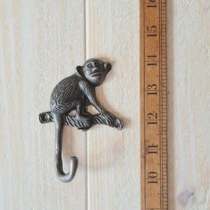 May include: A cast iron monkey hook in a dark grey finish. The monkey is perched on a branch, with its tail forming the hook. The hook is mounted on a light-coloured wooden surface. The hook measures approximately 13 cm in length.
