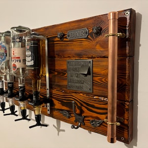 Op de afbeelding: Een rustieke houten bar display met vier drankfles dispensers. Het hout is gebeitst in een warme bruine kleur met een koperen pijp en messing accenten. Het bord luidt "BAR" en "ALL BEER MUST BE DRUNK IN THE MESS ROOM PROVIDED".