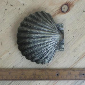 May include: A decorative, antique-style seashell-shaped object with a metallic finish. The shell has detailed ridges and a textured surface. A wooden ruler with inch markings is visible in the image.