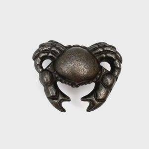 May include: A dark metallic, gunmetal-toned crab-shaped decorative object. The crab features a textured, rounded central shell and two prominent, articulated claws that curve inward towards the centre, creating a symmetrical, solid metal form.