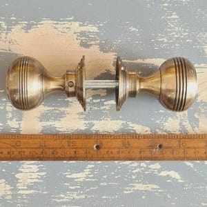 May include: A pair of brass door knobs with a ribbed, spherical design connected by a central metal spindle, positioned above a wooden ruler that shows the assembly measures approximately 16.5 cm in total length from end to end.