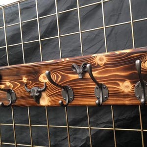 May include: A rectangular wall-mounted coat rack featuring a dark, flame-torched wood grain finish. The board is fitted with five metal hooks, interspersed with two decorative metal ram head sculptures and one central bull skull sculpture.