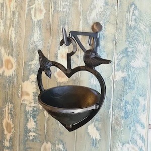 May include: A black metal wall planter shaped like a heart, with two bird figurines perched on the heart's frame. The planter has a bowl-shaped container for holding plants or other items. The wall mount is attached to a weathered wooden surface.