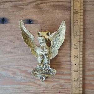 May include: A brass eagle door knocker with outstretched wings. The knocker is mounted on a wooden surface. A wooden ruler is next to the knocker, measuring approximately 15 cm in height.