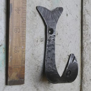 May include: A hand-forged, dark metal hook shaped like a fish. The hook features a tail, body, and a curved hook end. The hook is mounted on a light-coloured surface next to a wooden ruler.