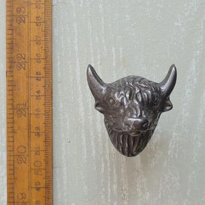 May include: A dark metallic wall hook shaped like a bull head with textured fur details and curved horns. A wooden ruler is positioned vertically on the left side, showing measurements in centimetres, with the bull head spanning roughly 5 centimetres.