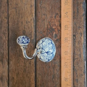 May include: A wall hook featuring a white ceramic knob and base with a blue floral pattern and a silver-toned metal arm. A wooden ruler is positioned next to the hook, showing the base has a diameter of approximately 5 cm.