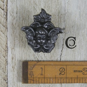 May include: A dark grey metal cabinet knob featuring a sculpted cherub face with wings and a floral crest at the top. The knob is positioned above a wooden ruler, showing the object is approximately 4.5 cm wide at its widest point.