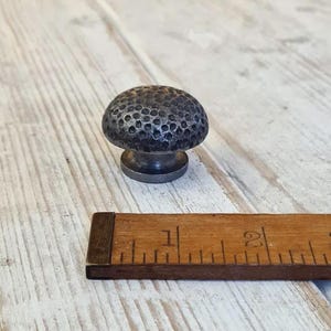 May include: A dark grey metal cabinet knob with a textured, dimpled surface. The knob is mushroom-shaped with a rounded top and a cylindrical base. A wooden ruler is positioned in the foreground, showing the knob is approximately 3.2 centimetres wide.