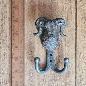 May include: A cast iron wall hook featuring a ram's head design with two hooks below. The hook is approximately 15 cm tall and has a rustic, antique finish. The ram's head has detailed features and curved horns.
