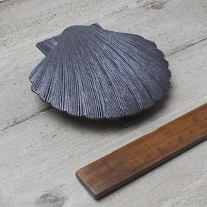 May include: A dark grey, cast iron seashell-shaped decorative object. The shell has detailed ridges and a textured surface. A wooden ruler is next to the shell. The object is likely a decorative element for home decor.