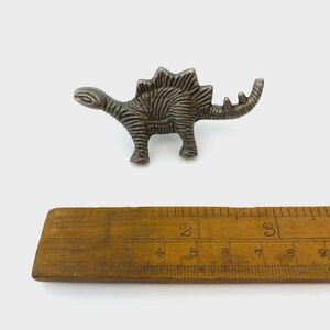 May include: A metallic, dark grey Stegosaurus-shaped knob or decorative piece with textured, ridged detailing along its back and body. It is positioned above a wooden ruler, showing the dinosaur figure measures approximately 7.6 cm in length.