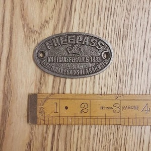 May include: Oval, cast metal Free Pass sign with the text "FREEPASS", "NOT TRANSFERABLE 1893", and "GREAT INDIAN PENINSULA RAILWAYS". The sign is approximately 5 cm long. A wooden ruler is in the foreground.