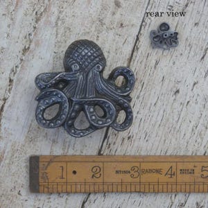 May include: A dark grey metal octopus-shaped cabinet knob with a textured head and curled tentacles. A smaller rear view of the knob is visible in the upper right corner. A wooden ruler shows the knob is approximately 6.5 cm in width.