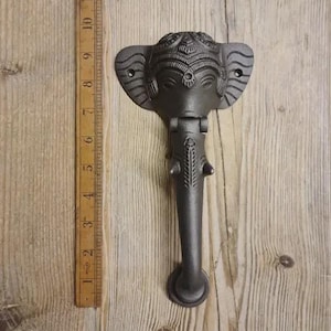 May include: A dark grey cast iron door knocker, shaped like an elephant's head with intricate carvings. The knocker is approximately 15 cm tall and is mounted on a wooden door.