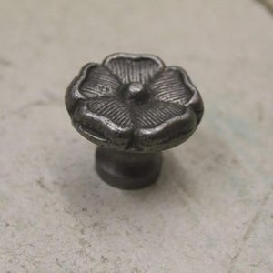 May include: A dark grey metal cabinet knob shaped like a five-petaled flower. The surface of the knob features a textured, ribbed pattern on each petal and a small, rounded centre point, providing a tactile and detailed appearance for furniture.