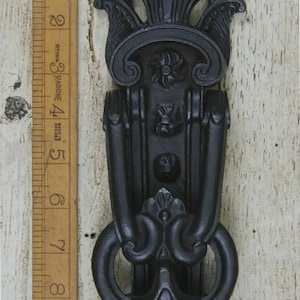 May include: Black cast iron door knocker with an ornate, symmetrical design. It features a decorative top, vertical bars, and a ring for knocking. The knocker is approximately 18 cm tall.