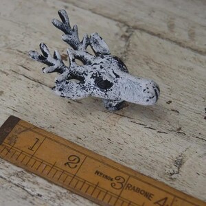 May include: A cast metal drawer knob shaped like a deer head with a distressed white and black finish. The knob is positioned next to a wooden ruler showing a length of approximately 7.5 cm, highlighting the scale of the deer-shaped hardware piece.