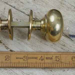 May include: A polished brass door knob set featuring two spherical knobs connected by a square spindle and two circular rosettes. A small plastic bag contains a black hex key and screws. A wooden ruler shows the set is approximately 12.7 cm long.
