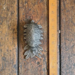 May include: A detailed, grey metal turtle-shaped door pull or cabinet knob. The decorative hardware is approximately 10 cm long and rests on a wooden surface with a ruler.