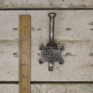 May include: A dark grey metal wall hook featuring the Coca-Cola logo embossed on the rectangular mounting plate. The hook has a vertical post and a curved lower prong. A wooden ruler is placed alongside, showing the hook is 15 cm in length.