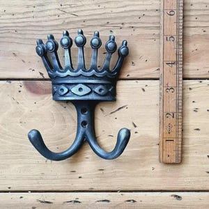 May include: A dark grey metal crown-shaped wall hook with two curved hooks. The crown features a decorative design with rounded details. A wooden ruler is next to the hook, showing measurements in centimetres.