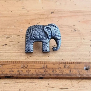 May include: A small, detailed elephant figurine with a metallic finish. The elephant has intricate patterns on its body and is on a wooden surface. The figurine is approximately 4 cm long.
