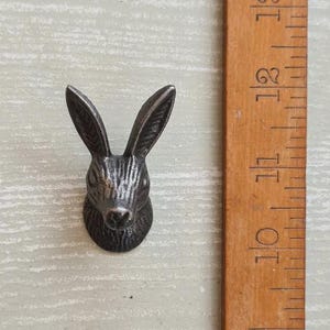 May include: A dark grey metal rabbit head wall hook with textured fur detailing and long upright ears. The rabbit head is positioned next to a wooden ruler showing measurements in centimetres, with the rabbit head height spanning approximately 5 cm.