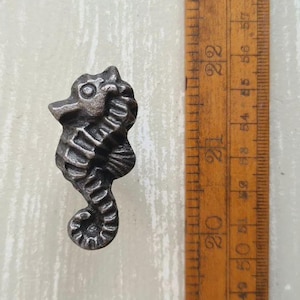 May include: A dark grey, metallic seahorse-shaped cabinet knob with a textured, ridged body. The knob is positioned next to a wooden ruler, showing the seahorse measures approximately 6.5 cm in height from the top of the head to the tail.