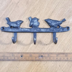 May include: A dark grey metal wall hook with three bird figures on a branch. The hook has three downward-facing hooks. The design includes ornate detailing, suitable for a rustic or nature-themed decor. The hook is approximately 40 cm in length.