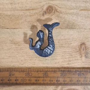 May include: A dark grey cast iron mermaid wall hook featuring a textured scale pattern on the tail. The mermaid figure is positioned above a wooden ruler that shows measurements in centimetres, with the hook spanning approximately 7.5 cm in height.