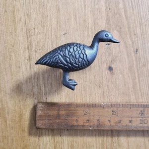 May include: A cast iron duck ornament with a textured body and a dark finish. The duck is standing on one leg. A wooden ruler is next to the duck, showing measurements in inches and centimetres.