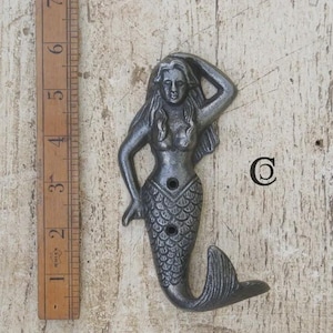 May include: A dark grey cast metal wall hook shaped like a mermaid with one arm raised to her head and a scaled tail. The hook measures approximately 18 cm in height when placed next to a wooden ruler for scale, featuring two mounting holes.