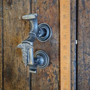 May include: A silver-toned, cast metal door knocker featuring a medical caduceus symbol. The knocker is mounted on a wooden door. A wooden ruler is placed next to the knocker, measuring up to 43 cm.