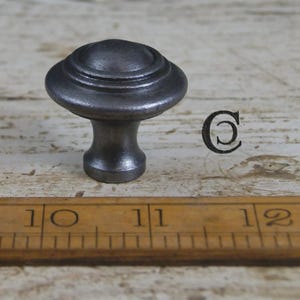 May include: A dark grey metal cabinet knob with a rounded top and a flared base. The knob is positioned above a wooden ruler showing measurements in inches, with the number 11 clearly visible. A copyright symbol is printed to the right of the knob.